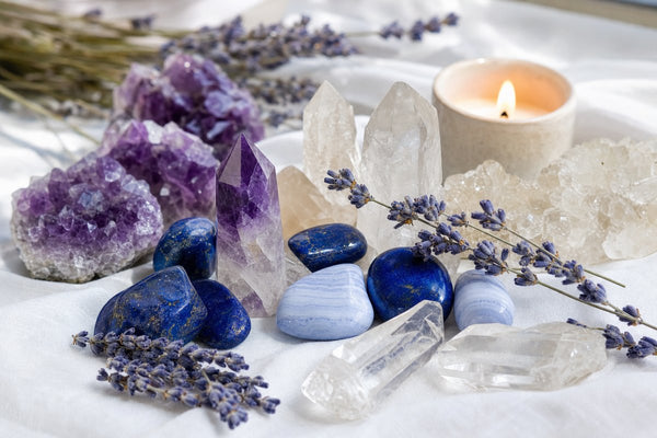 Amethyst clusters, lapis lazuli, clear quartz points, and blue lace agate crystals arranged on white linen with lavender and a candle